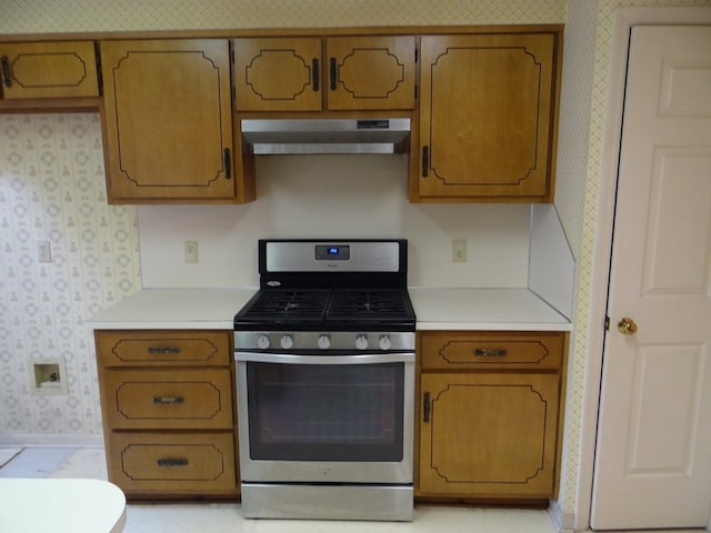 kitchen with ventilation hood, brown cabinetry, wallpapered walls, stainless steel range with gas stovetop, and light countertops