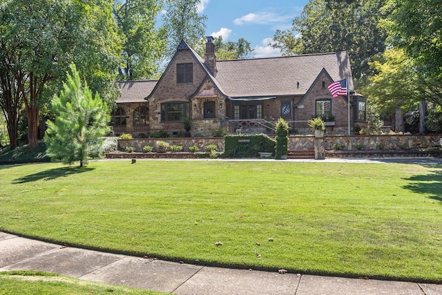 tudor house featuring stone siding, a chimney, covered porch, a front yard, and brick siding