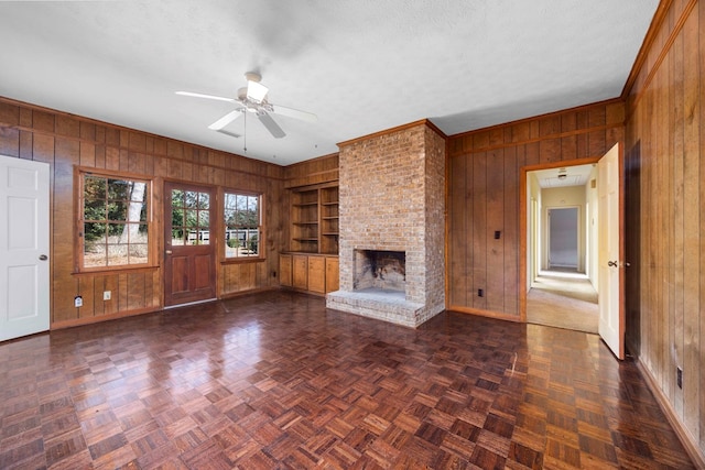 unfurnished living room with ceiling fan, a fireplace, a textured ceiling, built in shelves, and wood walls