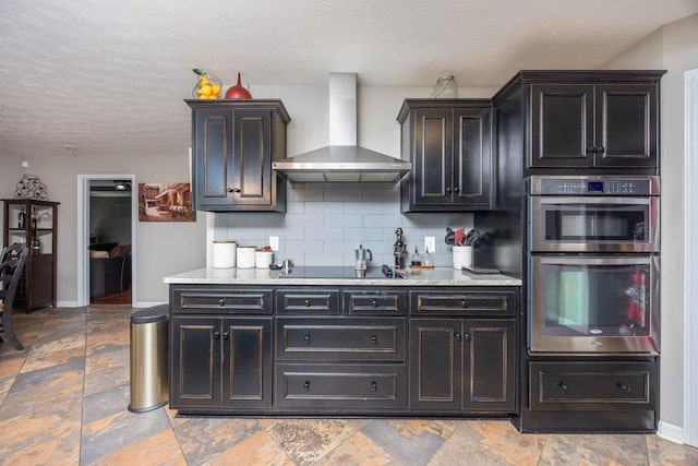kitchen with stone finish floor, tasteful backsplash, baseboards, double oven, and wall chimney exhaust hood