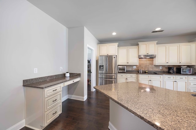 kitchen featuring dark stone counters, stainless steel fridge with ice dispenser, built in desk, under cabinet range hood, and tasteful backsplash