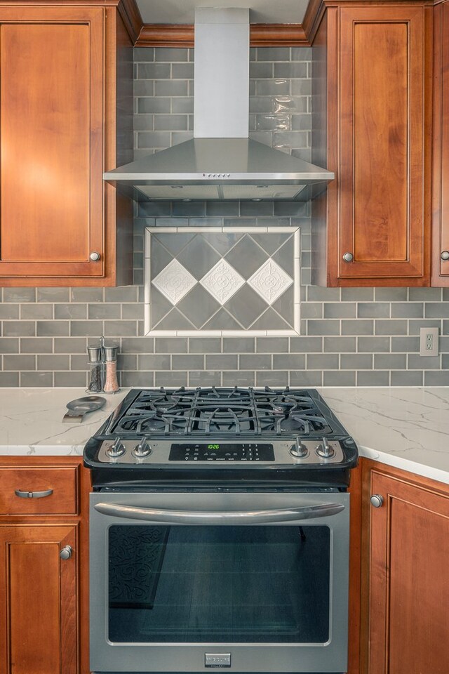 kitchen featuring stainless steel gas range, light stone countertops, brown cabinets, wall chimney exhaust hood, and backsplash