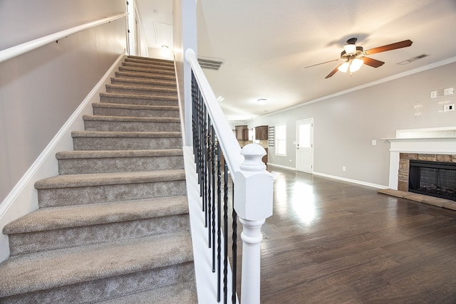 stairs featuring hardwood / wood-style flooring, ceiling fan, a stone fireplace, and ornamental molding