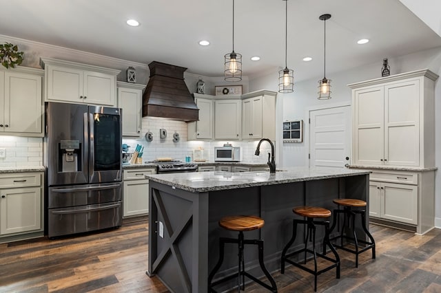 kitchen with sink, premium range hood, appliances with stainless steel finishes, light stone counters, and a center island with sink