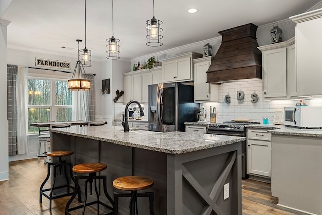 kitchen featuring appliances with stainless steel finishes, light stone countertops, a center island with sink, decorative light fixtures, and custom exhaust hood