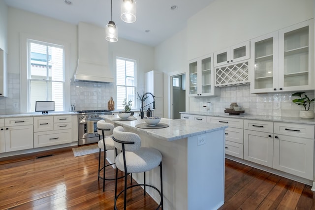 kitchen with backsplash, premium range hood, a center island with sink, stainless steel stove, and dark hardwood / wood-style floors