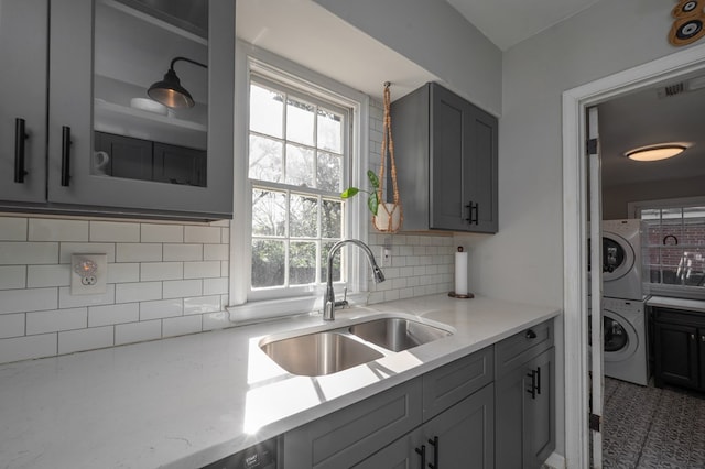 kitchen with backsplash, gray cabinets, stacked washer / drying machine, and a sink