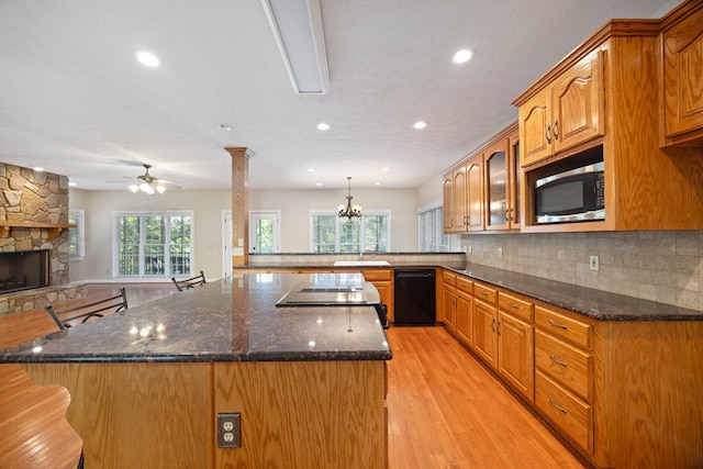 kitchen featuring black dishwasher, a stone fireplace, light hardwood / wood-style flooring, decorative columns, and kitchen peninsula