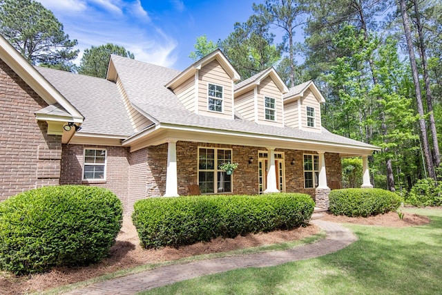 cape cod-style house with stone siding, a front yard, covered porch, and a shingled roof