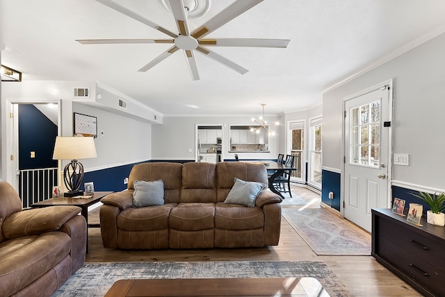 living room with light wood-type flooring, visible vents, crown molding, and ceiling fan with notable chandelier