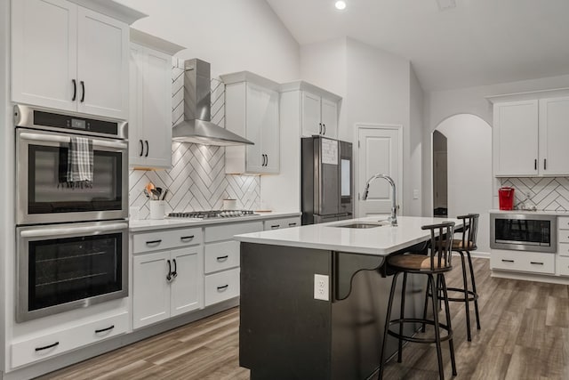 kitchen with wall chimney exhaust hood, stainless steel appliances, a kitchen island with sink, sink, and hardwood / wood-style floors