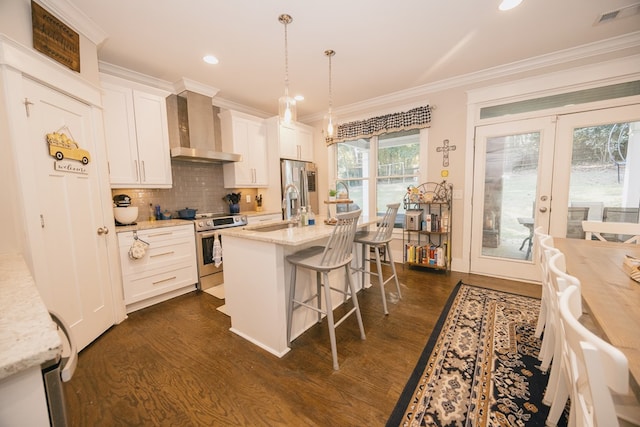 kitchen with stainless steel appliances, wall chimney range hood, decorative light fixtures, a kitchen island with sink, and white cabinets