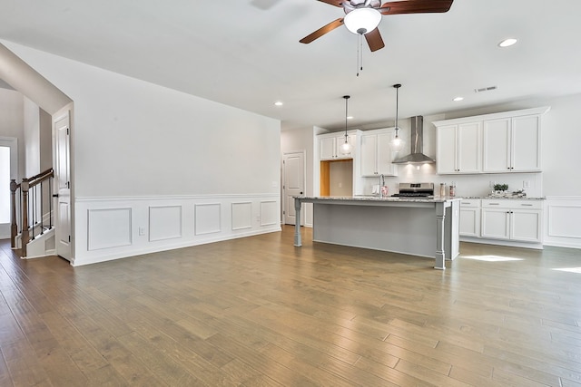 kitchen featuring a kitchen island with sink, hanging light fixtures, wall chimney exhaust hood, white cabinetry, and wood-type flooring