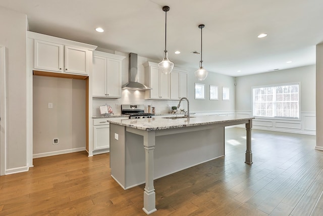 kitchen featuring wall chimney exhaust hood, stainless steel range, sink, white cabinetry, and an island with sink