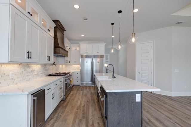 kitchen with premium range hood, premium appliances, pendant lighting, white cabinetry, and an island with sink