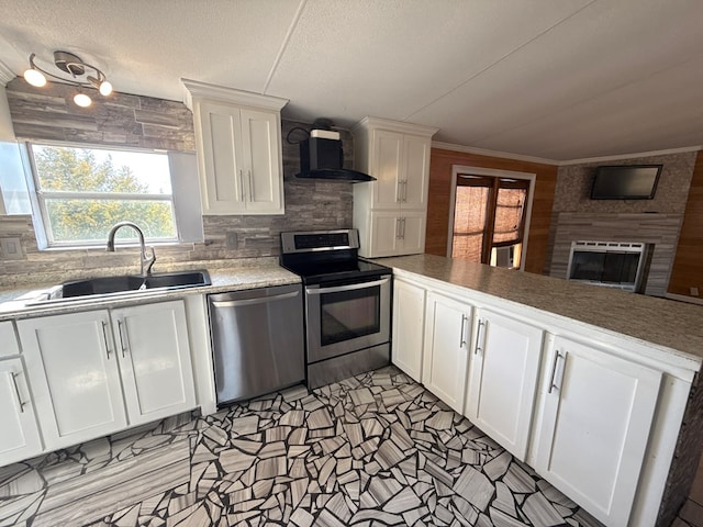 kitchen with white cabinets, a glass covered fireplace, wall chimney exhaust hood, stainless steel appliances, and a sink