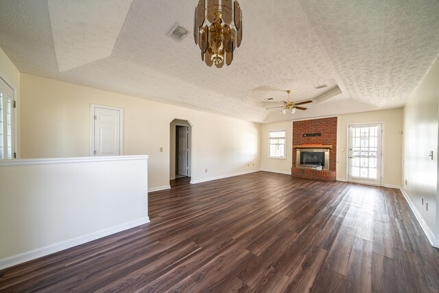 unfurnished living room featuring a brick fireplace, a textured ceiling, a tray ceiling, ceiling fan, and dark wood-type flooring