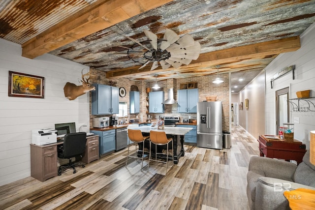 kitchen featuring blue cabinets, appliances with stainless steel finishes, wall chimney range hood, beam ceiling, and light wood finished floors