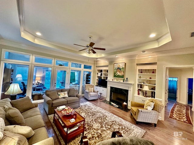 living room featuring ceiling fan, built in shelves, a raised ceiling, a high end fireplace, and wood-type flooring