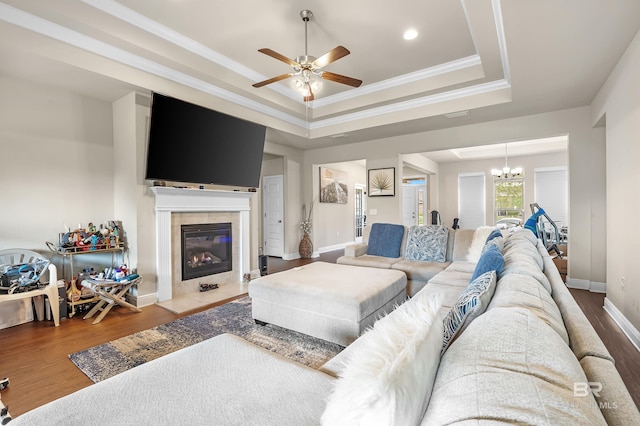 living room with a tile fireplace, ceiling fan with notable chandelier, wood-type flooring, a raised ceiling, and ornamental molding