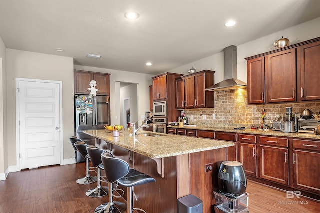 kitchen featuring a center island with sink, wood-type flooring, appliances with stainless steel finishes, sink, and wall chimney range hood