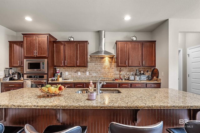 kitchen featuring light stone counters, sink, appliances with stainless steel finishes, a breakfast bar area, and wall chimney range hood