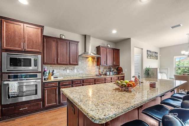 kitchen featuring light wood-type flooring, appliances with stainless steel finishes, light stone countertops, wall chimney exhaust hood, and a kitchen bar