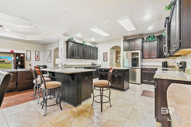 kitchen with a kitchen breakfast bar, light stone counters, dark brown cabinets, stainless steel appliances, and a kitchen island with sink