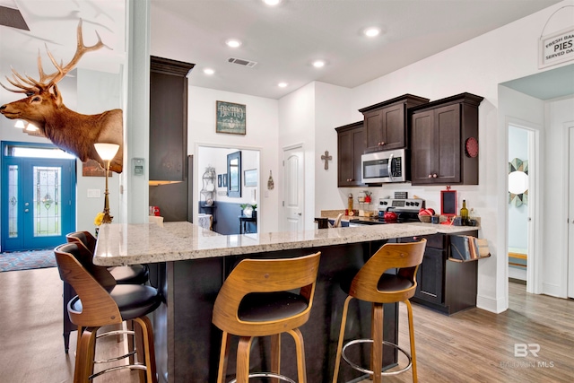 kitchen featuring a kitchen bar, stainless steel appliances, light stone counters, dark brown cabinetry, and light wood-type flooring