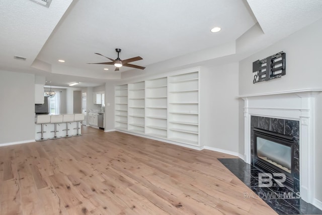 unfurnished living room with baseboards, a fireplace, a raised ceiling, light wood-type flooring, and ceiling fan with notable chandelier