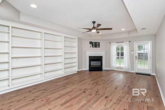 unfurnished living room featuring a tray ceiling, a fireplace, wood finished floors, baseboards, and ceiling fan