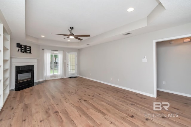 unfurnished living room featuring a raised ceiling, ceiling fan, built in features, light hardwood / wood-style flooring, and a tiled fireplace