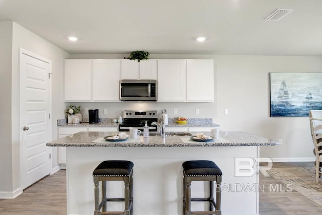 kitchen with a breakfast bar area, a kitchen island with sink, white cabinetry, stainless steel appliances, and light stone countertops