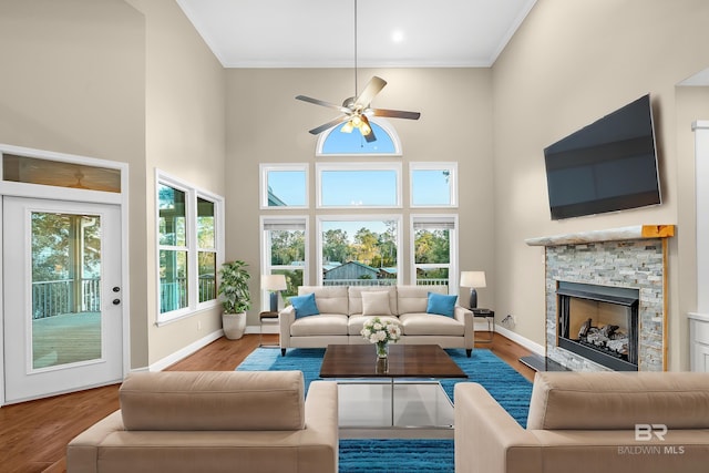 living room featuring wood finished floors, baseboards, a high ceiling, ornamental molding, and a stone fireplace