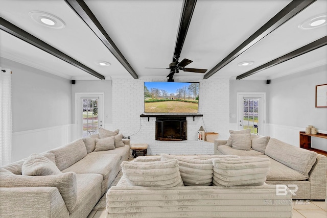 living room with ceiling fan, a wainscoted wall, brick wall, a fireplace, and beamed ceiling