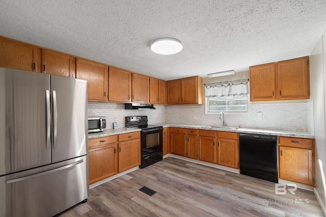 kitchen with brown cabinetry, a sink, under cabinet range hood, and black appliances