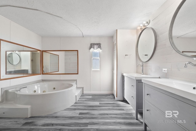 full bathroom with a textured ceiling, two vanities, a sink, wood finished floors, and a tub with jets