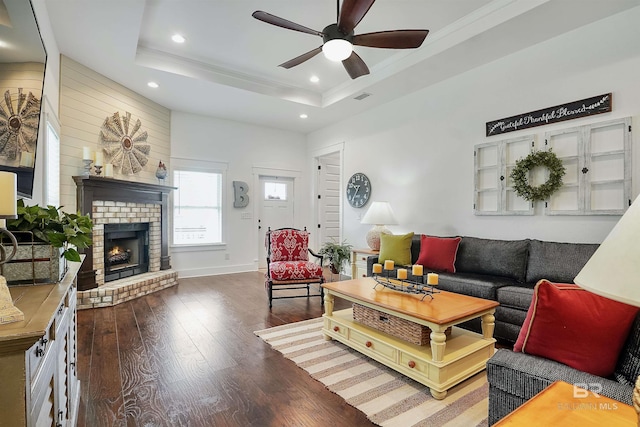 living room featuring a fireplace, ceiling fan, a tray ceiling, crown molding, and dark wood-type flooring