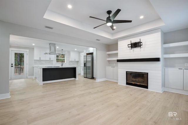 unfurnished living room with sink, ceiling fan, light hardwood / wood-style floors, a large fireplace, and a raised ceiling