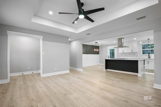 unfurnished living room featuring sink, ceiling fan with notable chandelier, a raised ceiling, and light wood-type flooring