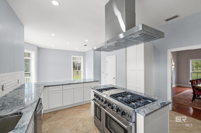 kitchen featuring decorative backsplash, island range hood, white cabinetry, light tile patterned flooring, and stainless steel appliances