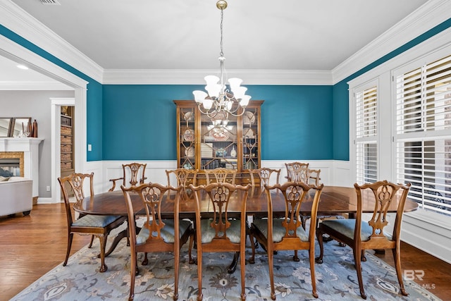 dining area featuring wainscoting, wood finished floors, crown molding, a fireplace, and a chandelier