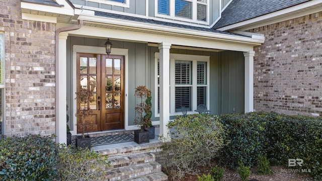 view of exterior entry with board and batten siding, french doors, brick siding, and roof with shingles