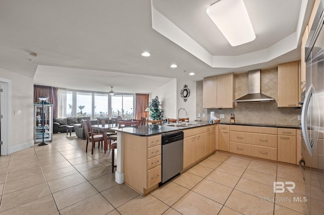 kitchen with wall chimney range hood, light brown cabinetry, open floor plan, dishwasher, and a sink