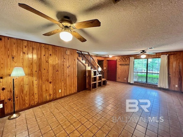 unfurnished living room with a textured ceiling, wood walls, a ceiling fan, baseboards, and stairway
