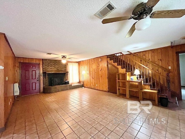 unfurnished living room featuring wooden walls, ceiling fan, visible vents, and stairs