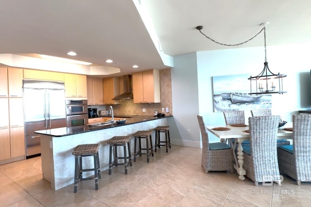 kitchen with a peninsula, appliances with stainless steel finishes, wall chimney range hood, light brown cabinetry, and dark countertops