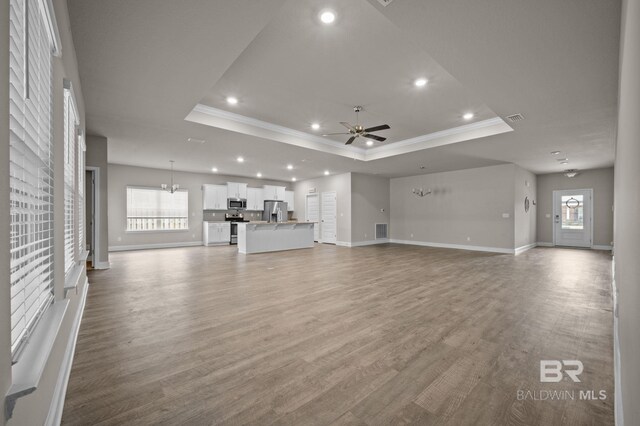 unfurnished living room with light hardwood / wood-style floors, a wealth of natural light, ceiling fan with notable chandelier, and a tray ceiling