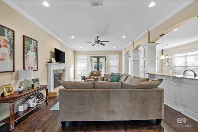 living room featuring ceiling fan with notable chandelier, crown molding, dark wood-type flooring, and a brick fireplace