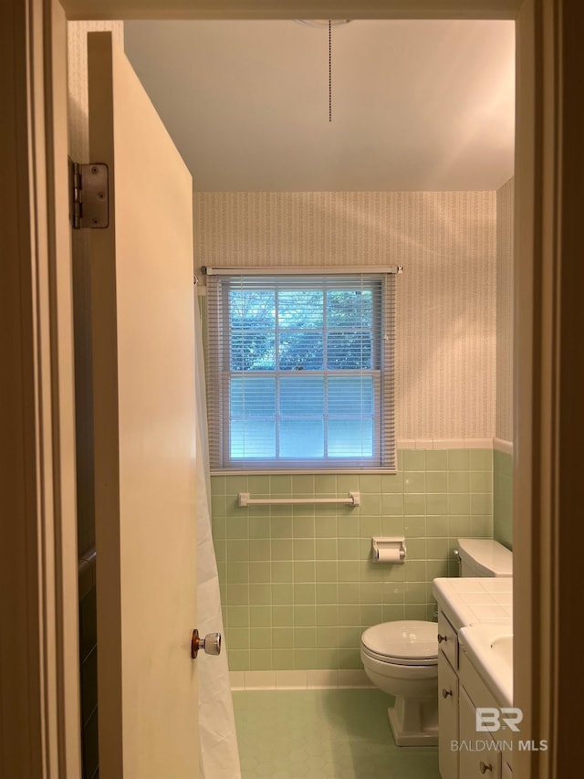 full bathroom featuring a wainscoted wall, tile walls, toilet, vanity, and wallpapered walls